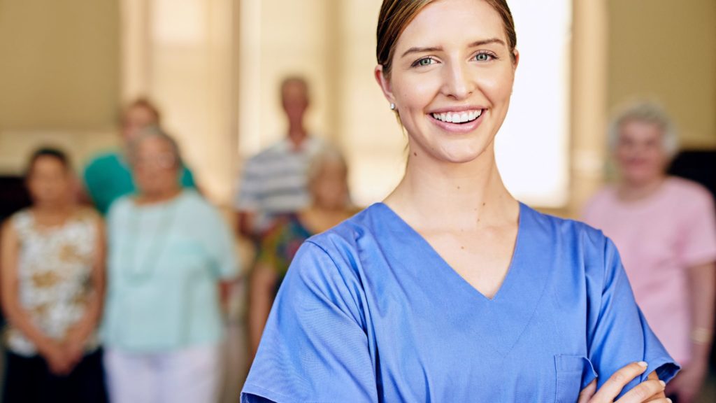 A caregiver stands in the foreground, with a senior figure appearing blurry in the background.