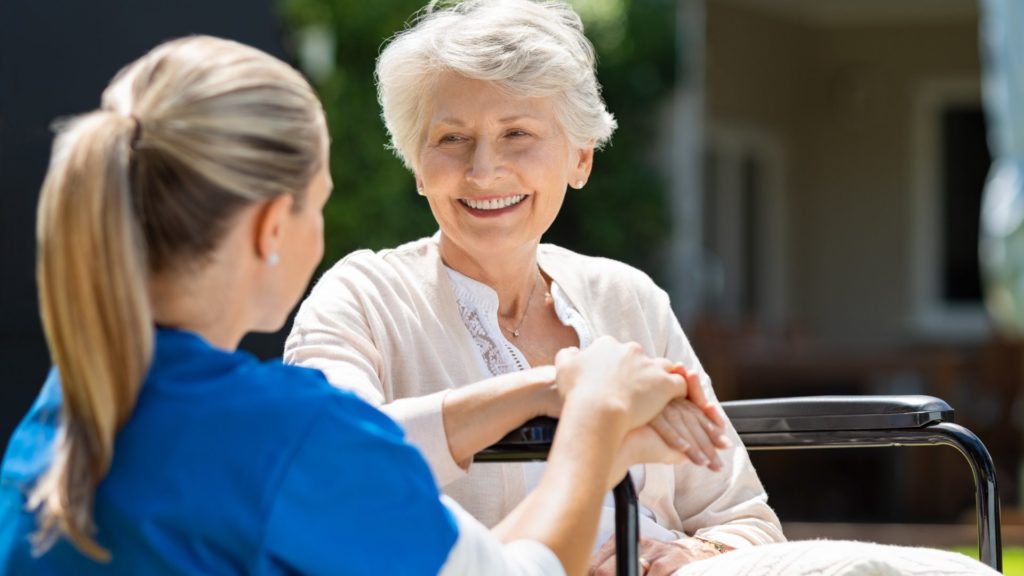 A senior sits in a chair beside a caregiver, who is gently holding their hand.